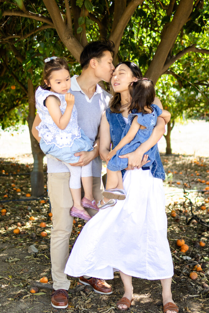 Dad kisses mom’s forehead while holding their daughters during a family photoshoot under the trees in the San Francisco Peninsula.
