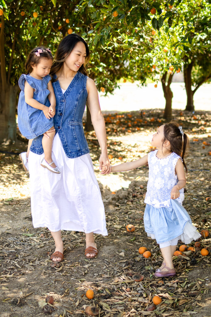 Mom in white dress and denim vest holds her toddler and walks hand in hand with her older daughter during a family photoshoot in a park