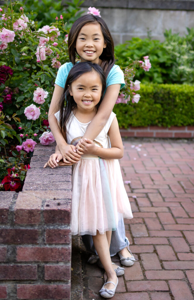 Sisters laughing while sitting on a brick ledge surrounded by blooming roses