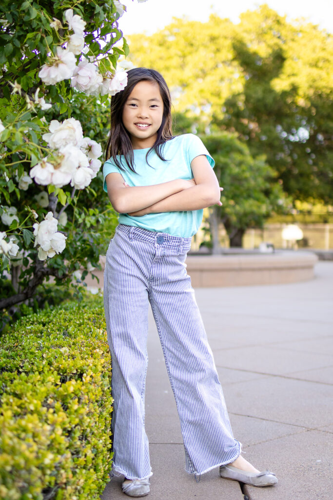 Confident young girl poses by blooming white roses with arms crossed