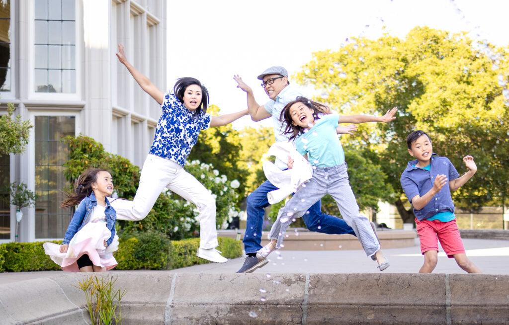 Family of five jumps midair with joyful expressions in front of elegant building