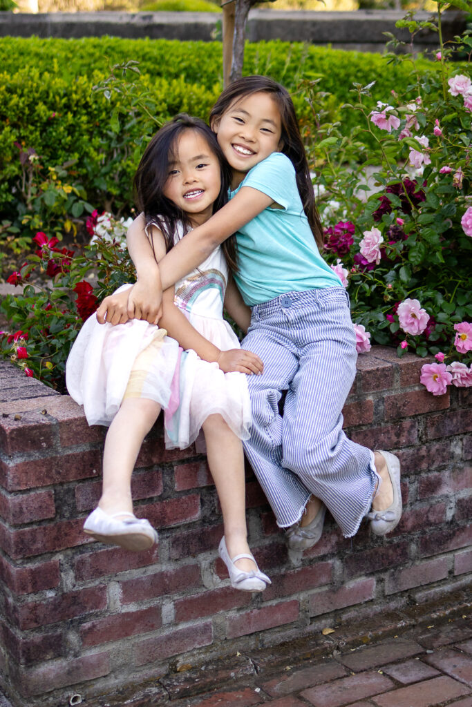 Sisters smiling and hugging beside a brick rose garden wall