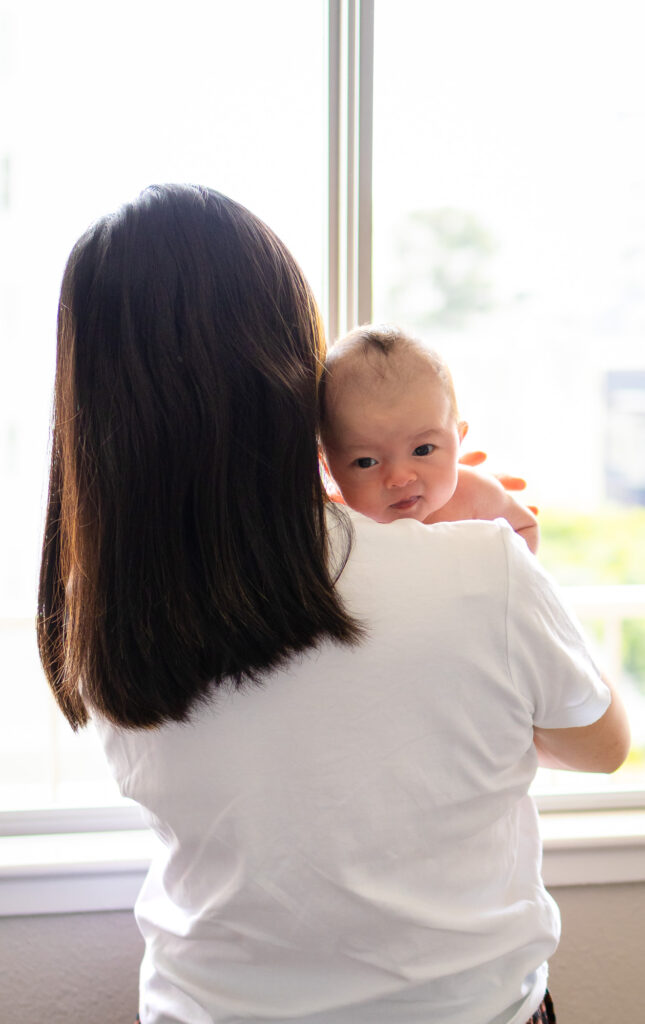 Mom holds baby over her shoulder near a bright window, capturing an everyday moment of calm