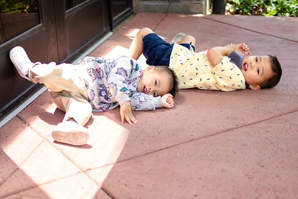 Two toddlers lying down and laughing on a patio during a playful family photoshoot