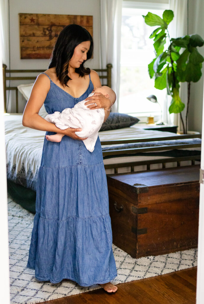 Mom standing barefoot in a bedroom holding her swaddled newborn, surrounded by peaceful home details for a best time for lifestyle newborn photos feature