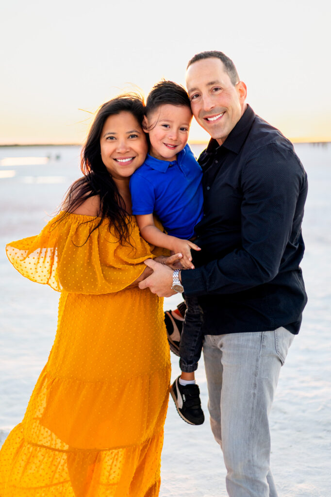 Smiling family of three poses together on the white salt flats at Alviso Marina County Park – Ellobelle Photography