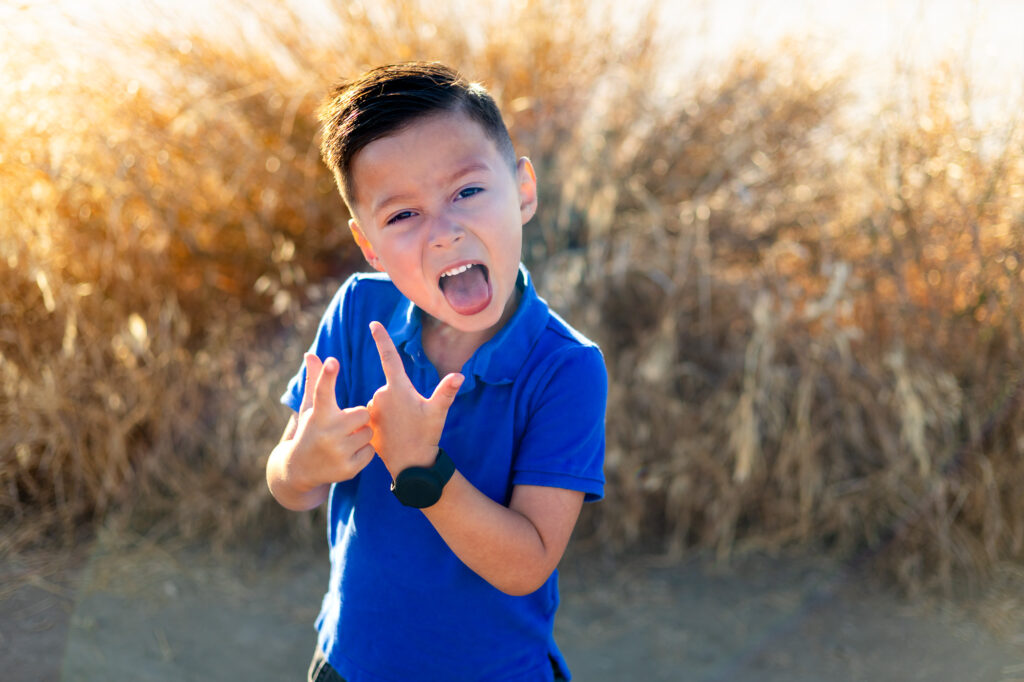 Boy sticks out tongue and pretends to shoot finger guns with dry grass in the background at Alviso Marina County Park – Ellobelle Photography