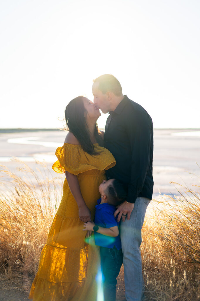 Couple shares a romantic kiss while their son hugs them from below at Alviso Marina County Park – Ellobelle Photography