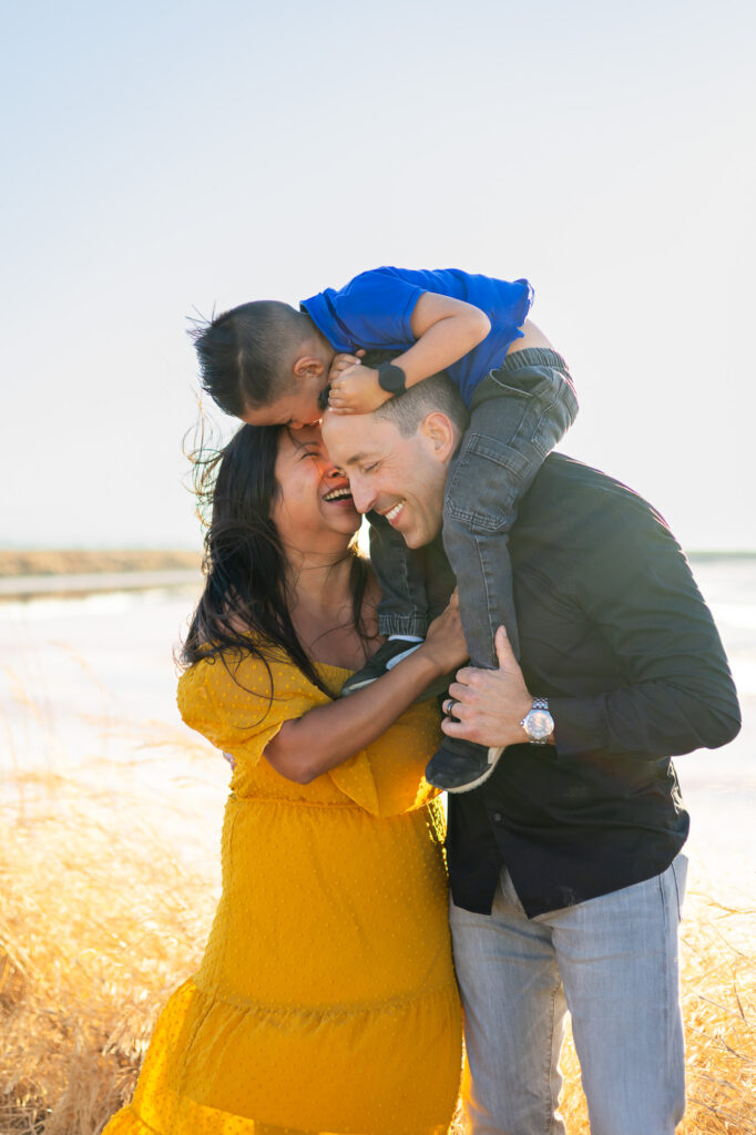 Young boy kisses mom while sitting on dad’s shoulders as the family laughs at Alviso Marina County Park – Ellobelle Photography