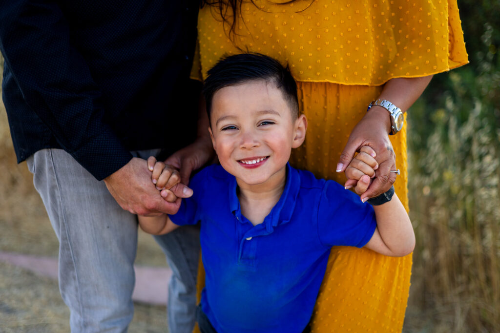 Young boy smiles while holding his parents’ hands during a warm sunset session at Alviso Marina County Park – Ellobelle Photography