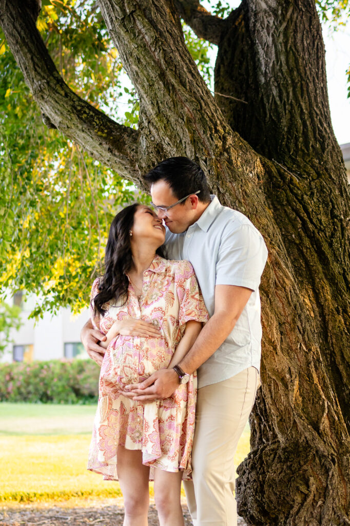 Couple smiling and touching foreheads under large tree during maternity session