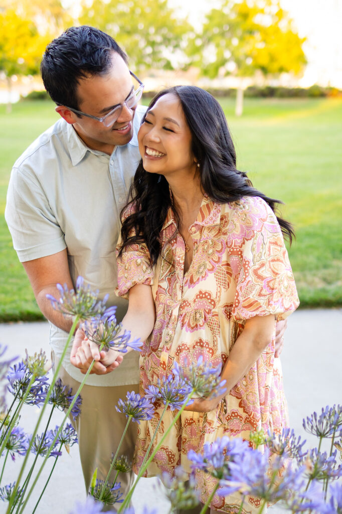 artner embraces pregnant woman as they smile and lean in among purple blooms