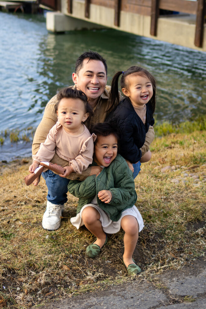 Dad and three daughters smiling by the water near the marina bridge at San Mateo County marina