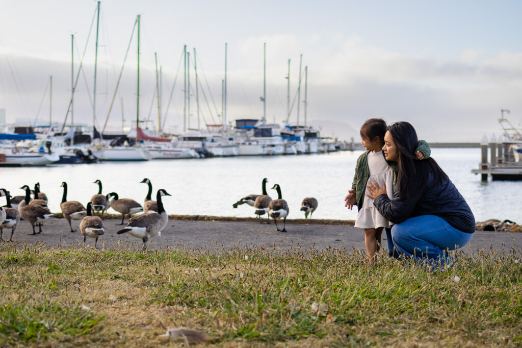 Mama and daughter watching geese by the water at Oyster Point waterfront with sailboats in the background