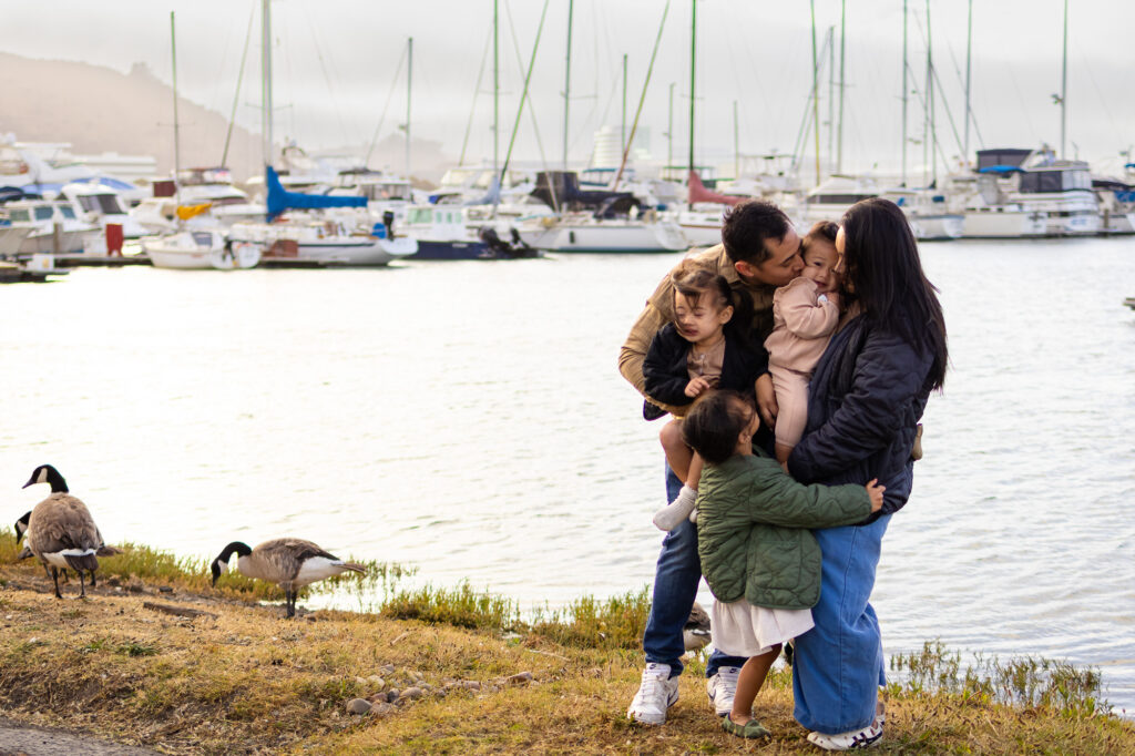 Family of five cuddling on the marina shore with geese and boats at Bay Area shoreline marina