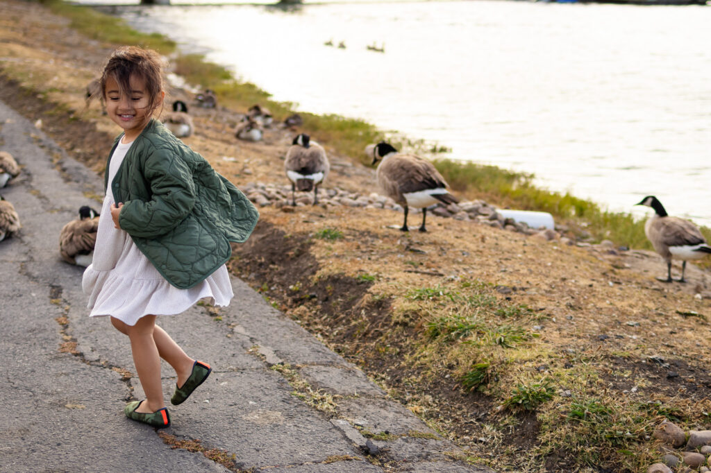 Young girl spinning joyfully near a flock of geese by the water at San Francisco Peninsula waterfront park