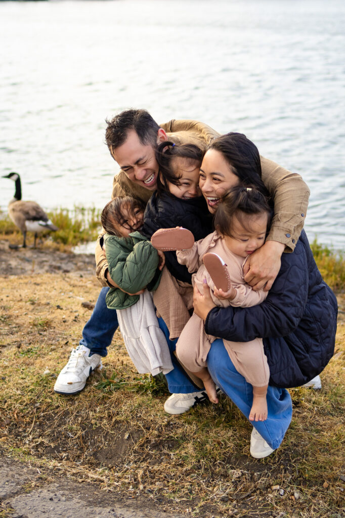 Family laughing and embracing near the shoreline with geese and calm waters at San Mateo County marina - Ellobelle Photography