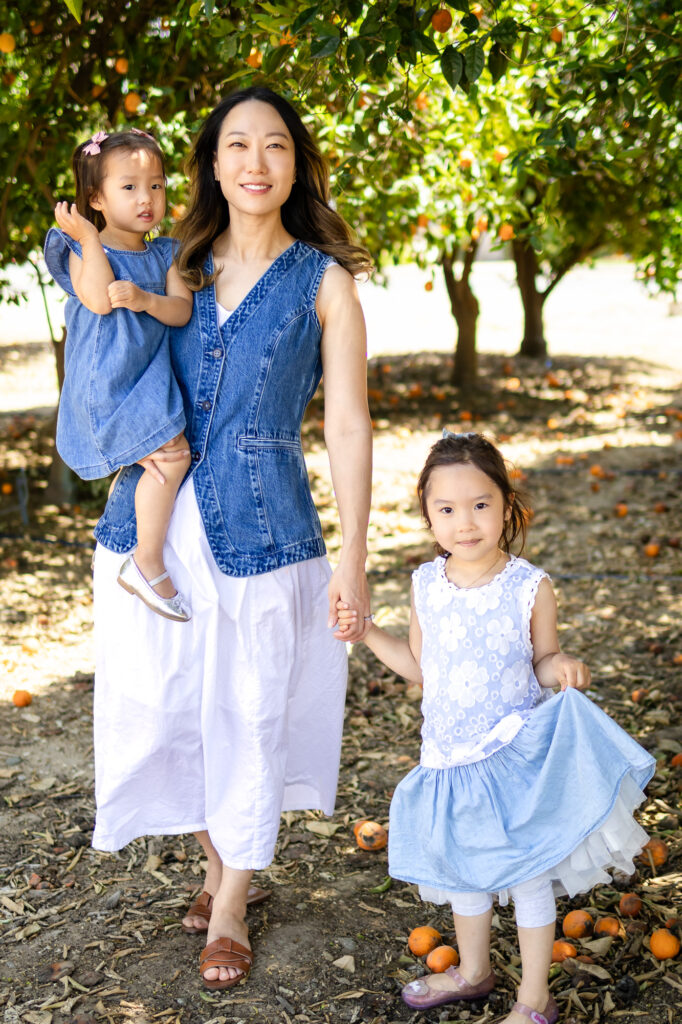 Mom in white dress and denim vest holding two daughters under an orange tree – What to Wear for Family Photoshoot