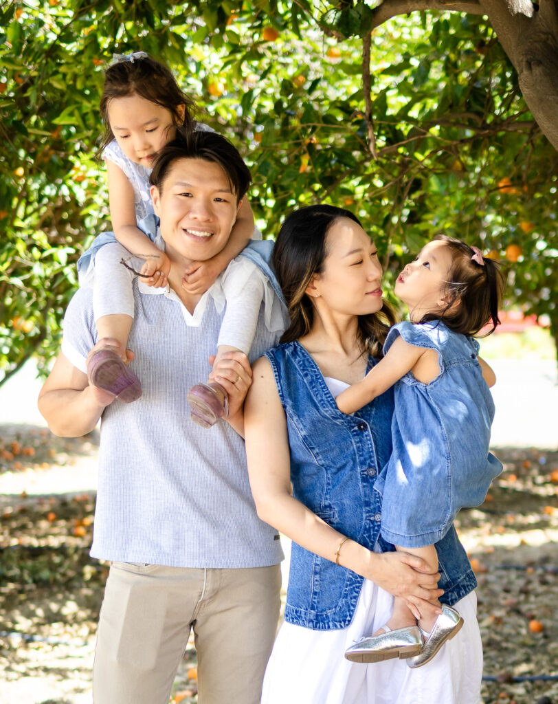 Candid moment of family with daughters on shoulders and in arms, smiling under orange trees