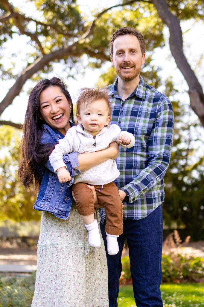 What to Wear for Family Photoshoot - Ellobelle Photography highlights beaming parents holding their baby, framed by golden trees.