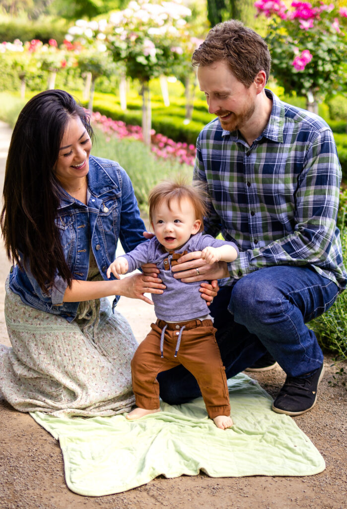 What to Wear for Family Photoshoot proud parents helping baby stand on blanket in blooming garden.