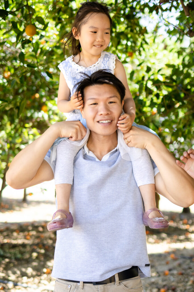 Daughter on dad’s shoulders smiling while walking through orchard – What to Wear for Family Photoshoot