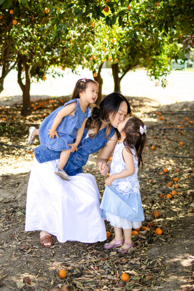 Mother crouching to kiss daughter, toddler in arms during orchard session
