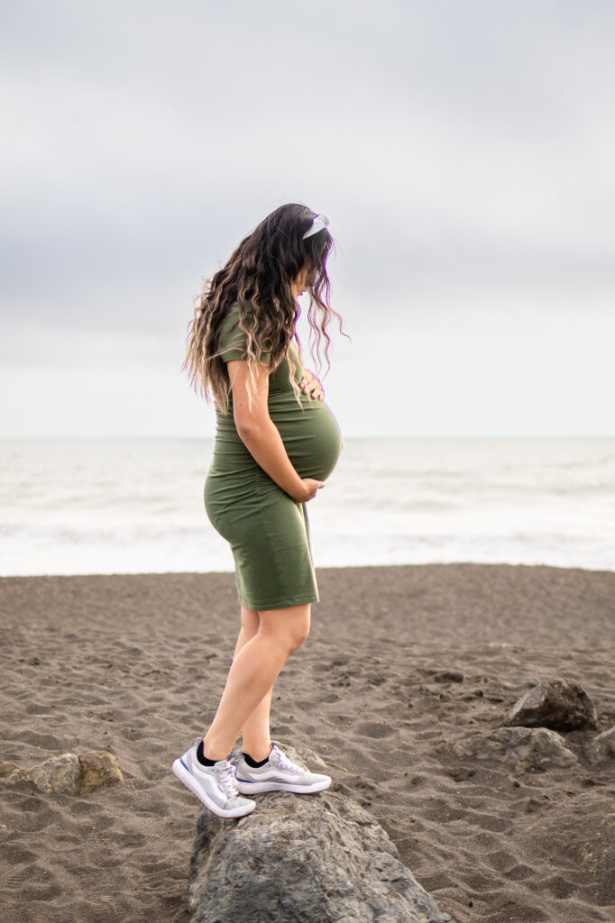 Pregnant mother standing on a rock at the beach during golden hour, highlighting the best time for maternity photos.