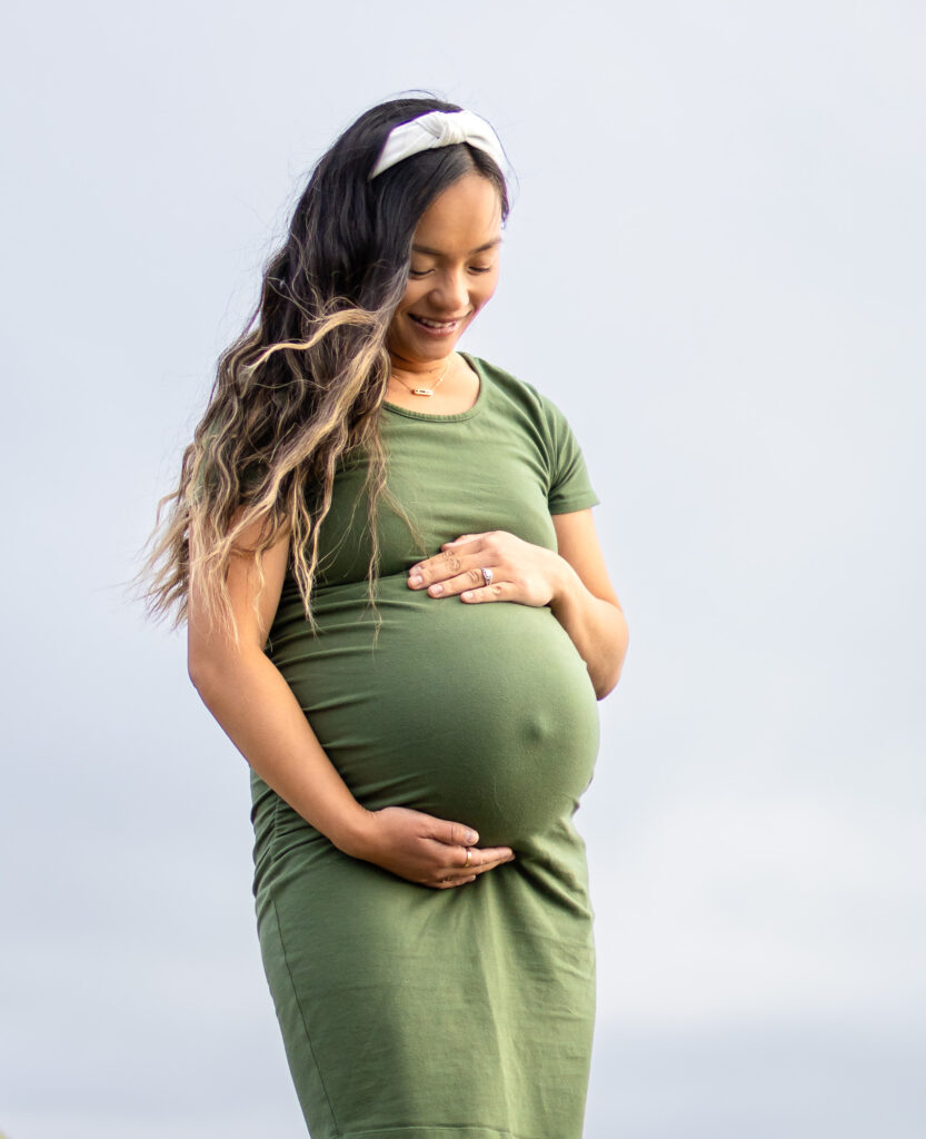 Pregnant woman smiling and holding her belly in a green dress during golden hour, capturing the best time for maternity photos.