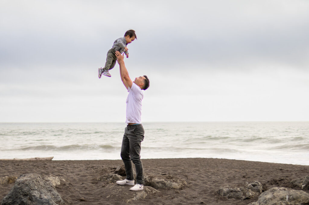 Dad lifting toddler high in the air on the beach with the ocean in the background during maternity session.
