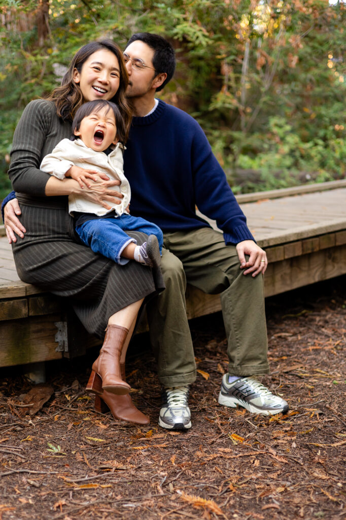 Joyful family moment with parents kissing child at Redwood Grove Los Altos – Ellobelle Photography
