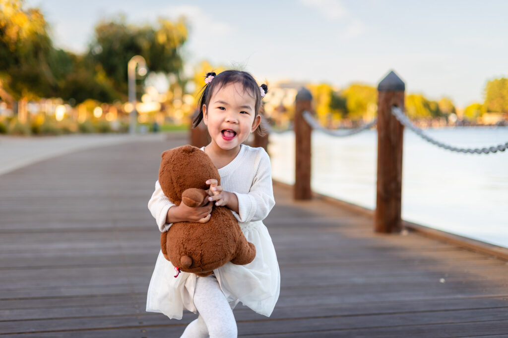 Little girl running with teddy bear along the boardwalk at grassy fields in Foster City