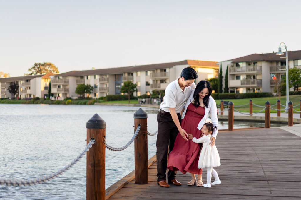 Parents hugging daughter close on the dock of candid moments in the grassy field at a Foster City park