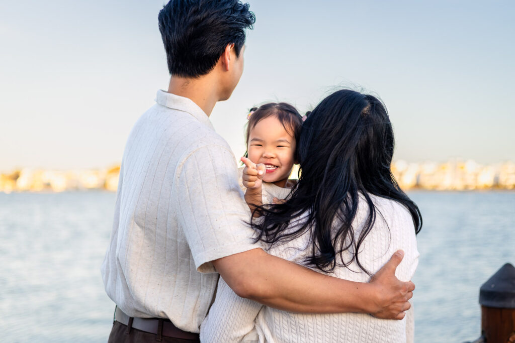 Daughter smiling while being held by parents near the lagoon at Leo Ryan Park