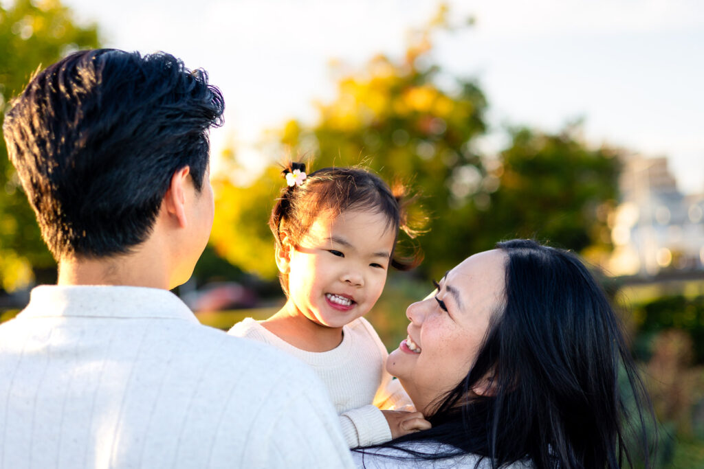 Toddler smiling in parents’ arms with golden trees in the background