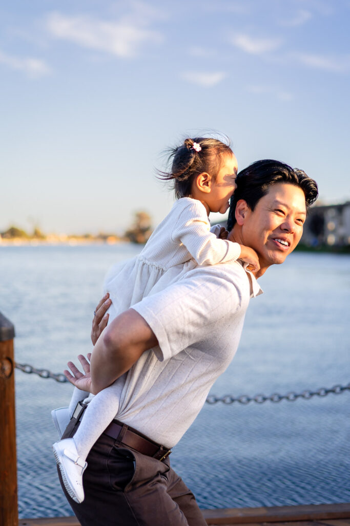 Daughter on dad’s back laughing near the water at tree lined walkways by the water