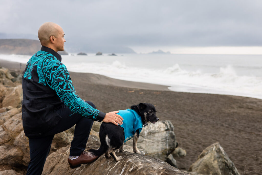 Man and dog sitting on coastal rocks looking out at the ocean during a reflective outdoor dog portraits in the Bay Area