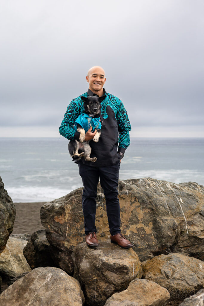 Full-body beach portrait of man holding his dog on rocky shore