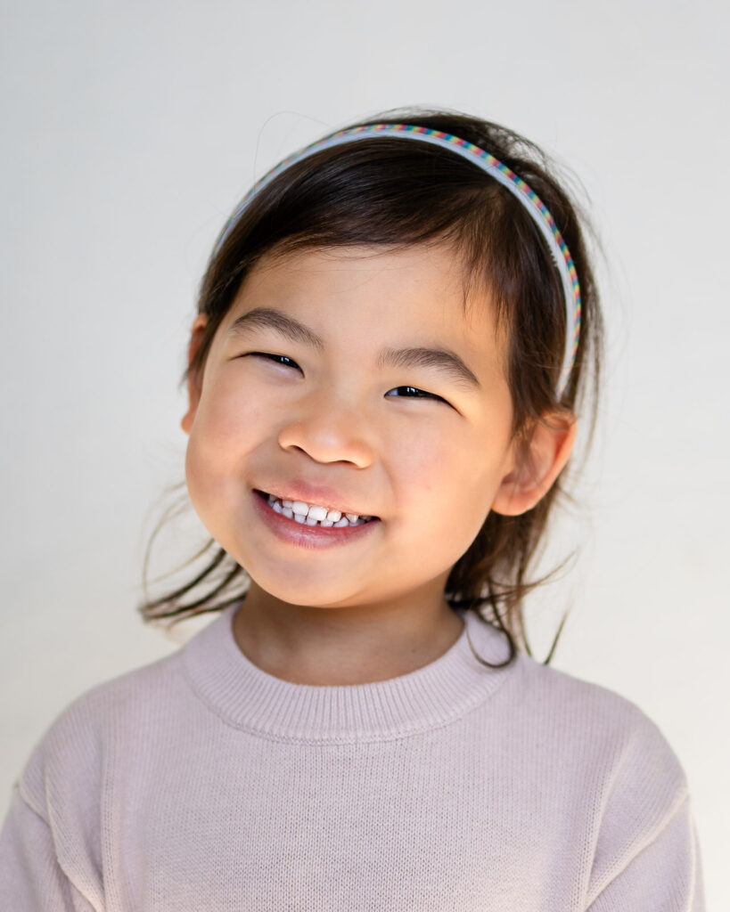 Young girl smiling in natural light against a light backdrop photographed by Bay Area Preschool Photographer in San Mateo – Ellobelle Photography