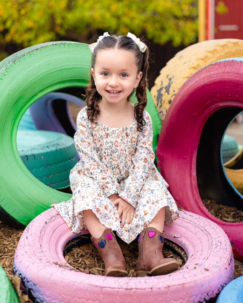 Preschool girl sitting on colorful playground tires during lifestyle portraits with Bay Area Preschool Photographer in San Bruno – Ellobelle Photography