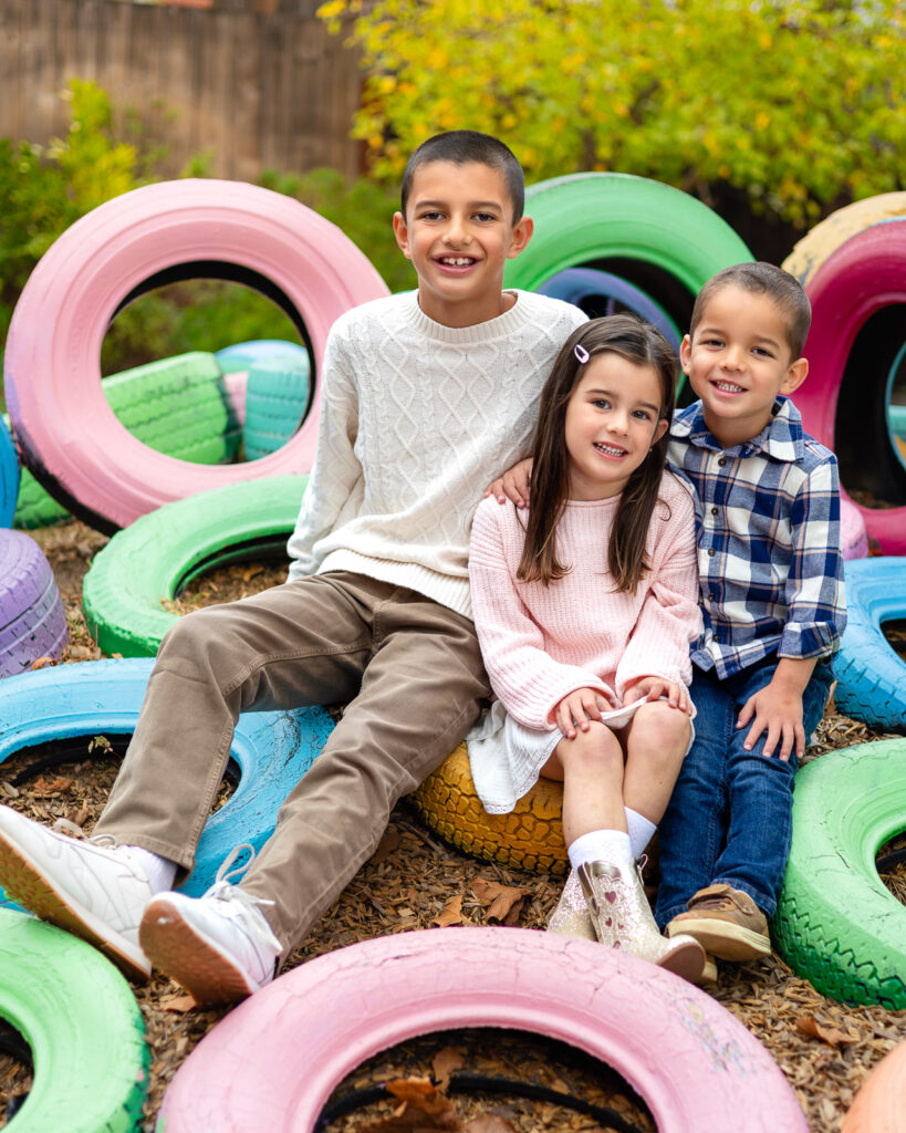 Three siblings sitting on colorful tires at Millbrae Nursery School during outdoor picture day playground portraits Ellobelle Photography