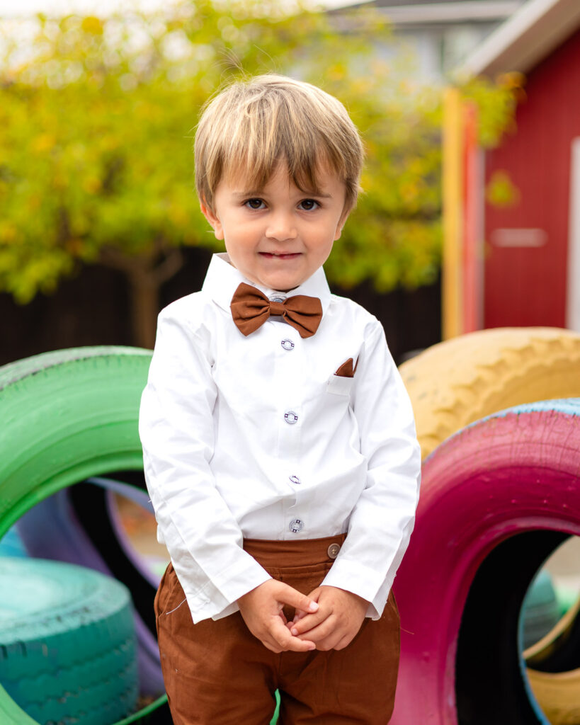 Young boy in a bow tie standing near colorful tire structures at Millbrae Nursery School Ellobelle Photography