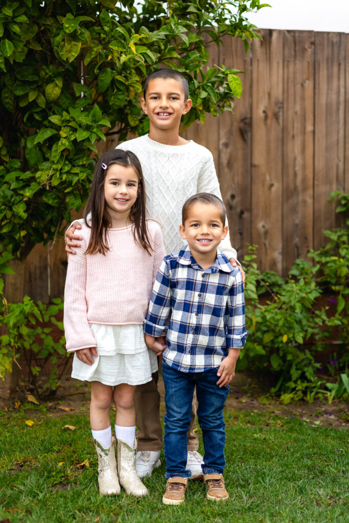 Three children smiling in the garden area at Millbrae Nursery School during school picture day Ellobelle Photography