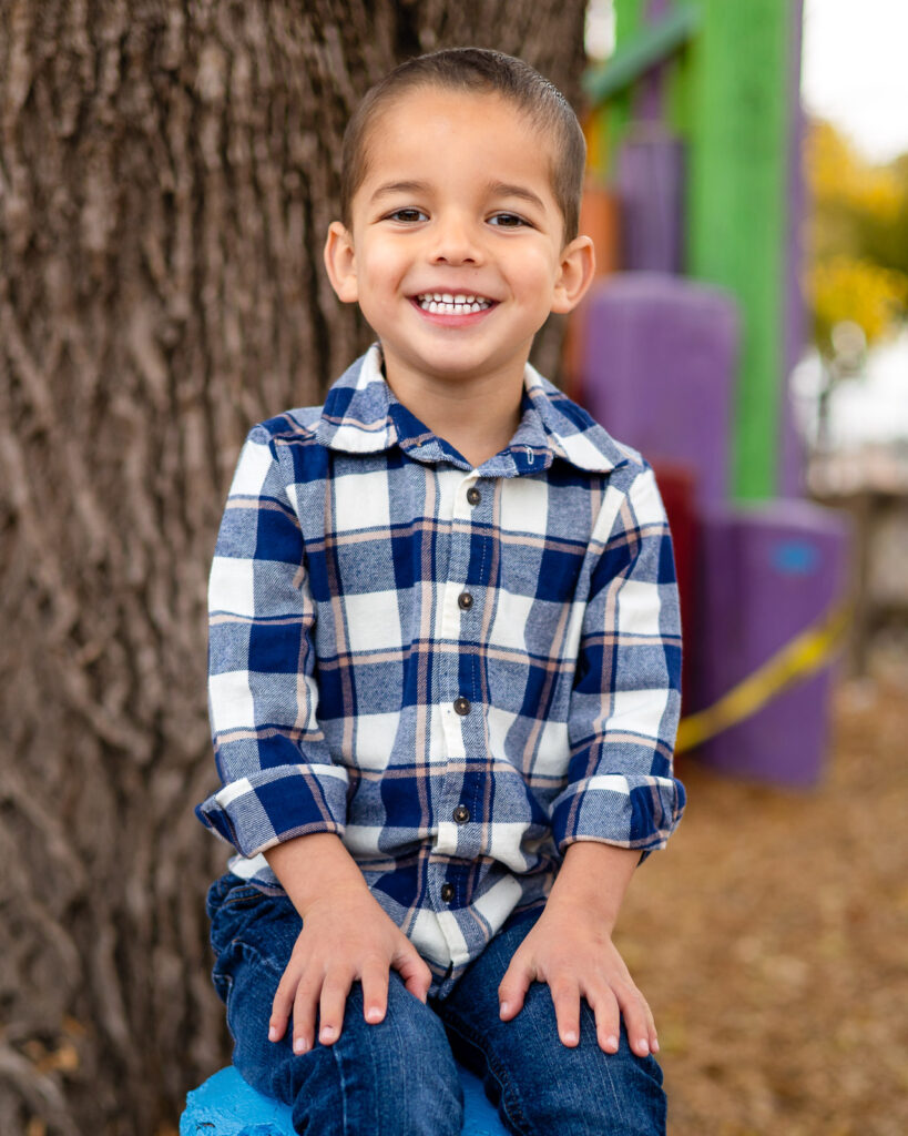 Young boy smiling while sitting on a painted tire near the large tree at Millbrae Nursery School Ellobelle Photography