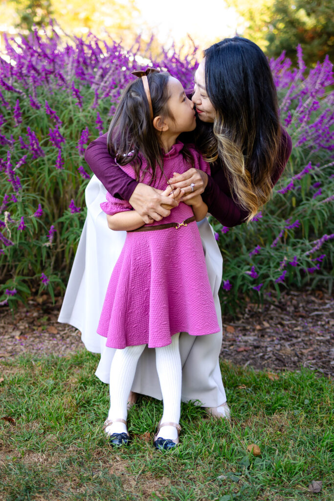 Mom and daughter kissing near purple flowers in a park for Family Photoshoot guides