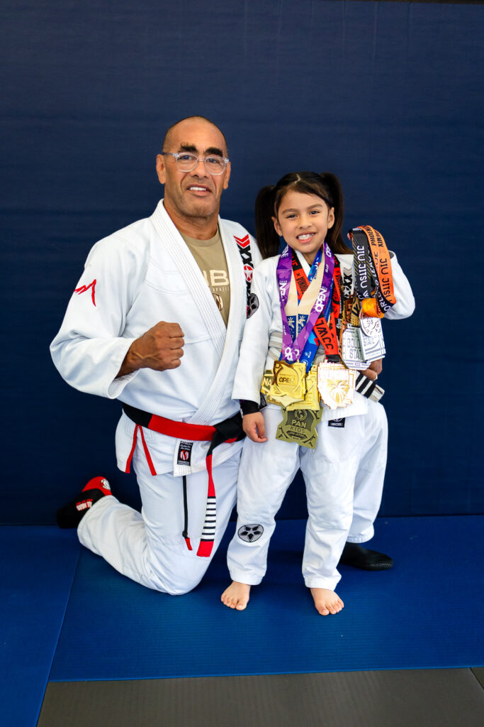 Coral Belt Marco Nascimento kneeling beside young athlete with medals at family friendly martial arts 
