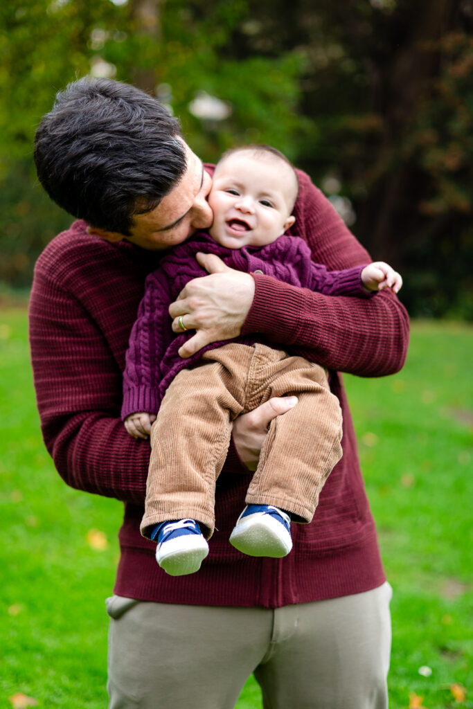 Dad kissing his smiling baby during a playful moment at candid moments under the trees in Millbrae