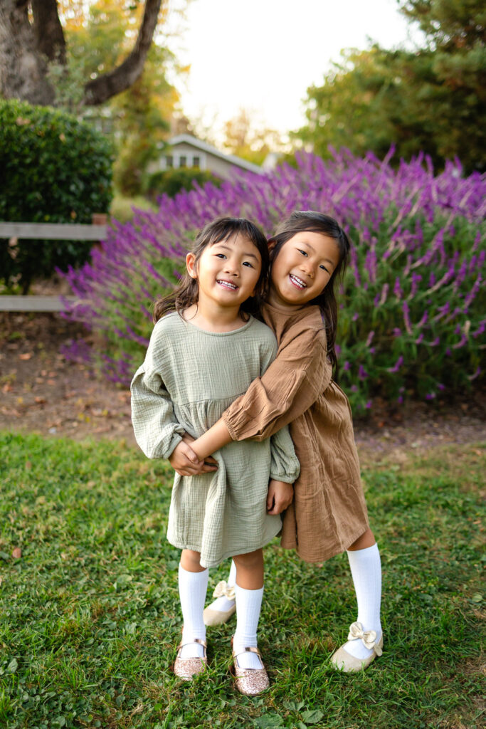Sisters hugging in front of purple flowers in a garden for tips for family photos with toddlers