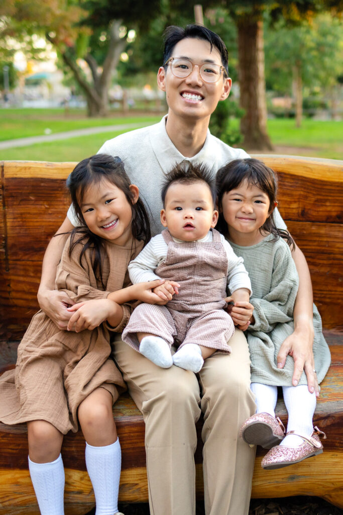Dad holding his three children on a wooden bench in a garden with children