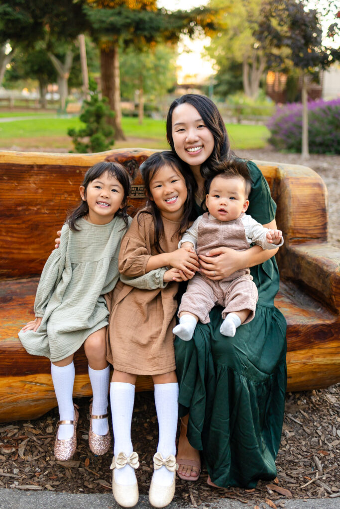 Mom holding her three kids on a wooden bench in a park with two kids and a baby